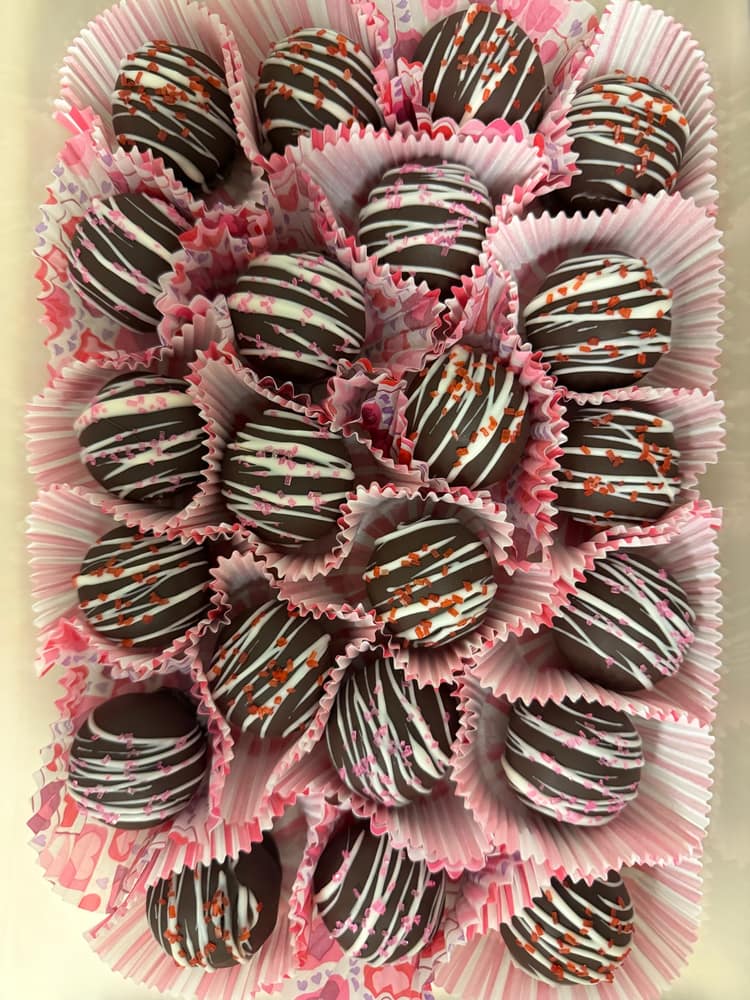 A tray of chocolate-dipped cake balls drizzled with white chocolate and topped with pink and red sprinkles, in pink patterned candy cups