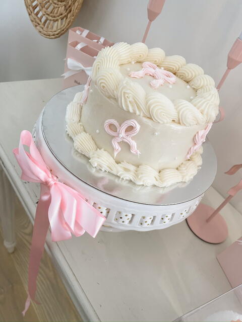 Small white buttercream cake with piped shell borders and pale-pink fondant bows, on a white pedestal stand