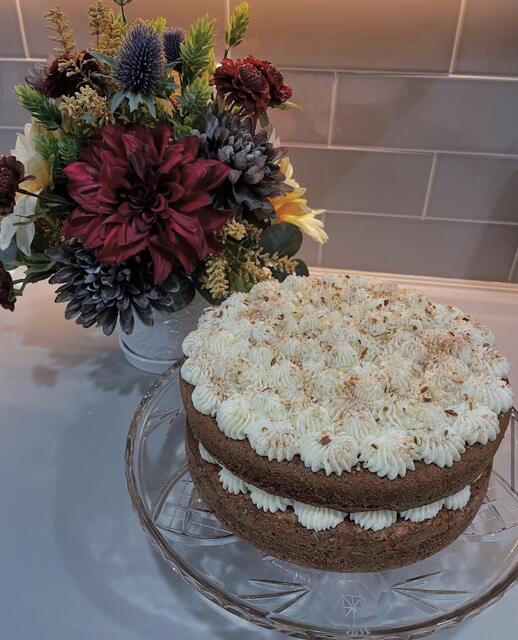 Two-layer chocolate cake with piped cream rosettes and toasted almond topping on a glass cake stand, next to a dark floral arrangement