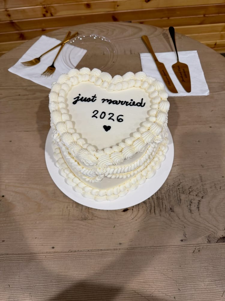 Heart-shaped white cake with vintage-style piped buttercream borders and hand-lettered 'just married 2026' in black icing, on a wood table