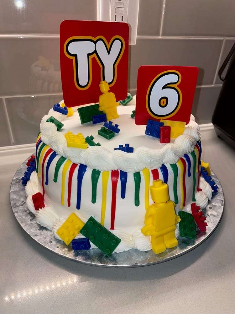 Round white birthday cake with a rainbow icing drip and candy building blocks, topped with red 'TY' and '6' signs and a yellow toy minifigure