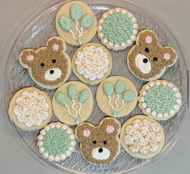 Decorated sugar cookies in soft green and white — teddy-bear faces, balloon bunches and piped florals — on a clear glass platter