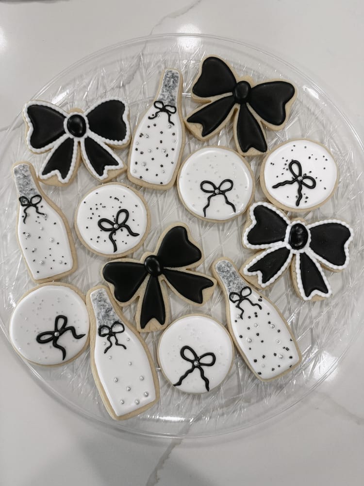 Black-and-white decorated sugar cookies on a clear glass plate: champagne-bottle shapes, bow shapes, and round cookies piped with black bows and silver dragees