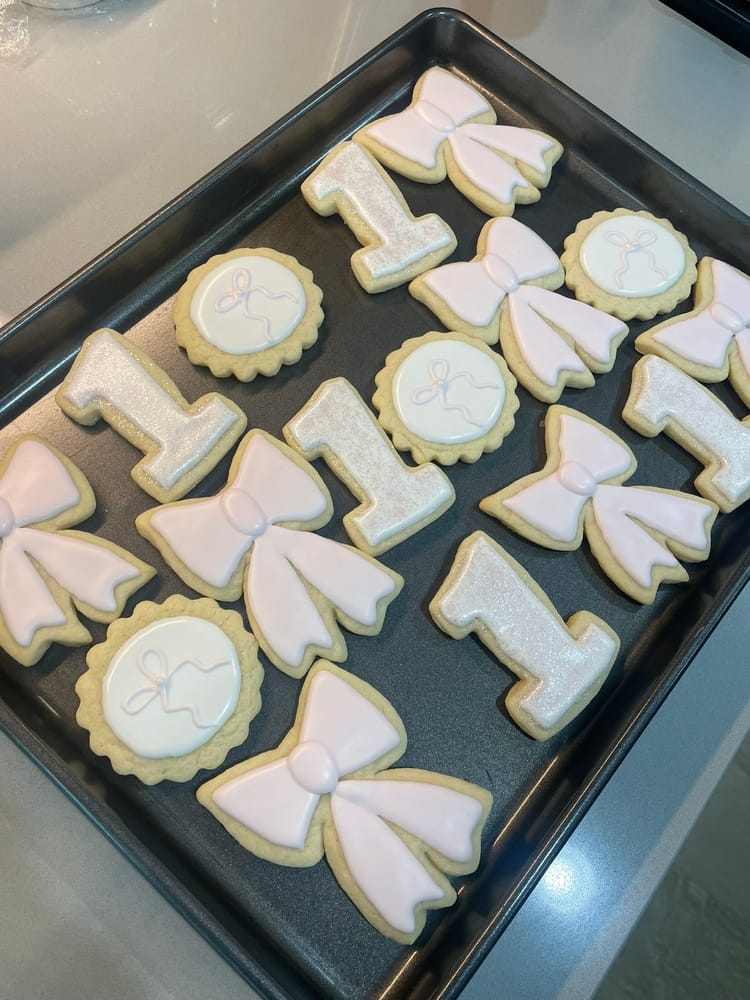 Pale-pink and white decorated sugar cookies on a baking sheet: glittered number-one shapes, bow shapes, and round cookies piped with small bows