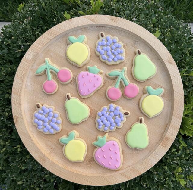 Pastel decorated sugar cookies shaped like strawberries, pears, cherries, lemons and grapes, arranged on a round wooden board