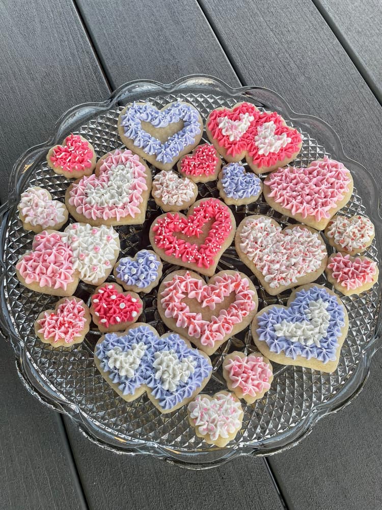 Heart-shaped sugar cookies piped with textured buttercream flowers in pink, rose, lavender and white, arranged on a cut-glass plate