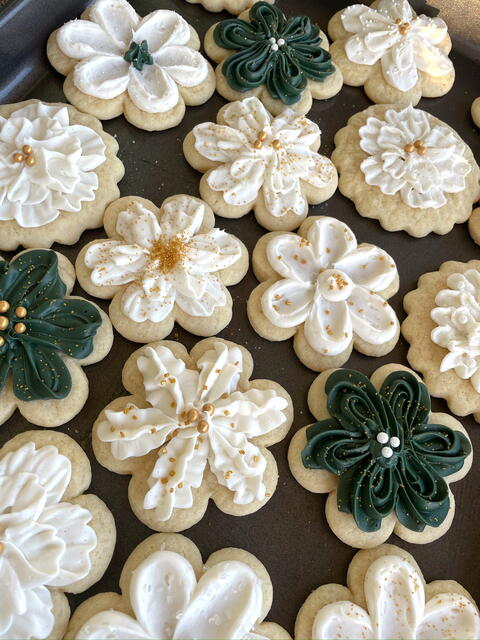 Flower-shaped sugar cookies iced with white and deep-green buttercream petals and gold sugar pearls, on a baking tray