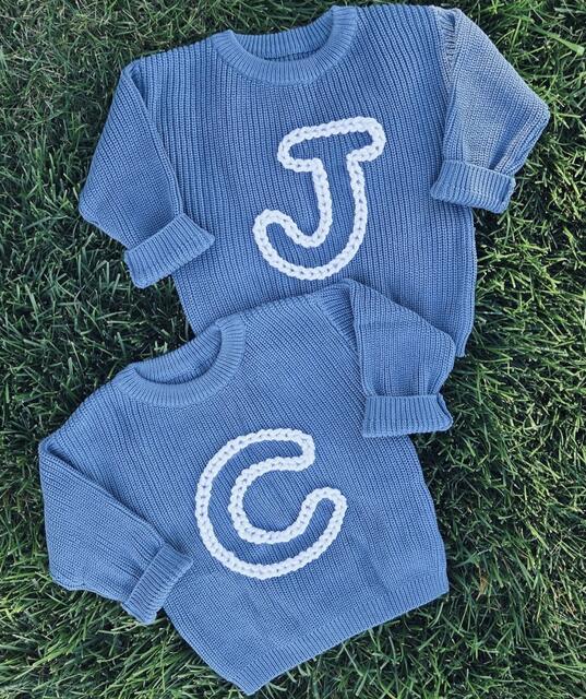 Two blue knit sweaters laid on grass, each hand-embroidered with a large single initial
