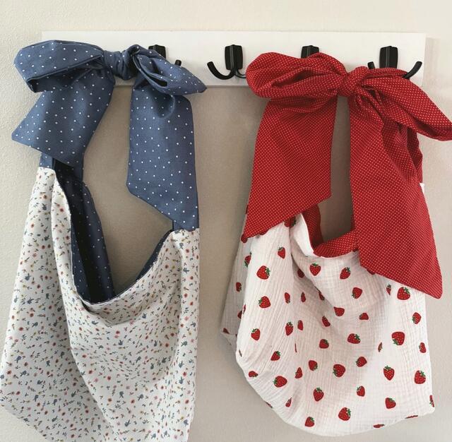 Two slouchy handmade tote bags hanging on wall hooks, one with a navy polka-dot fabric bow and floral-print body, the other with a red polka-dot bow and strawberry-print body